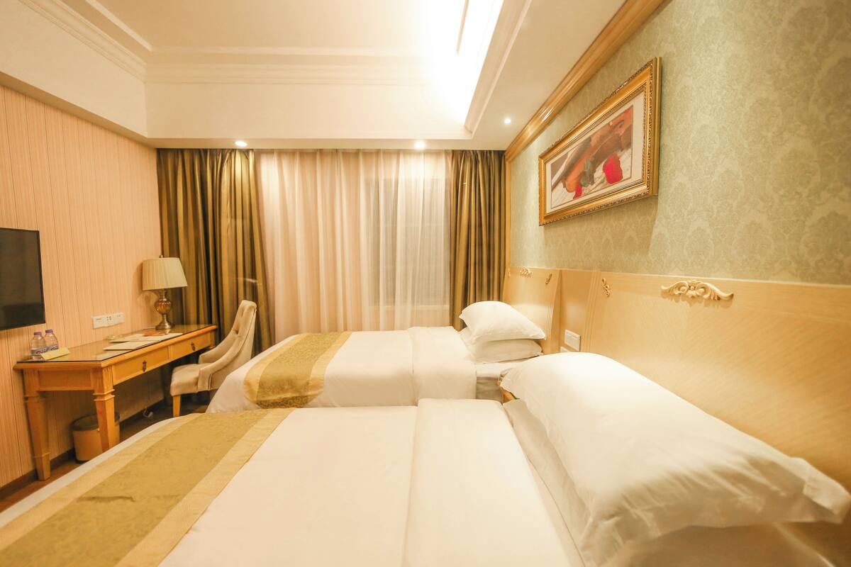A hotel room with two beds, a desk, a chair, a TV, and a painting on the wall.