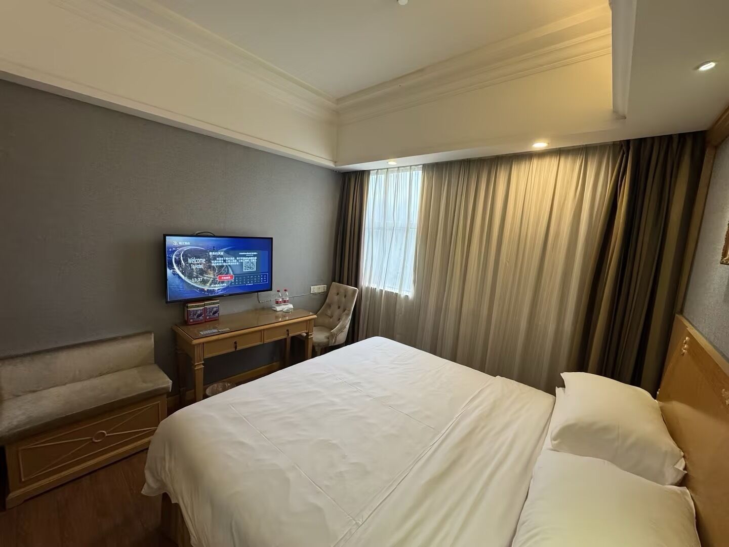 A hotel room with a bed, a TV on a stand, a desk with a chair, and a window with curtains.