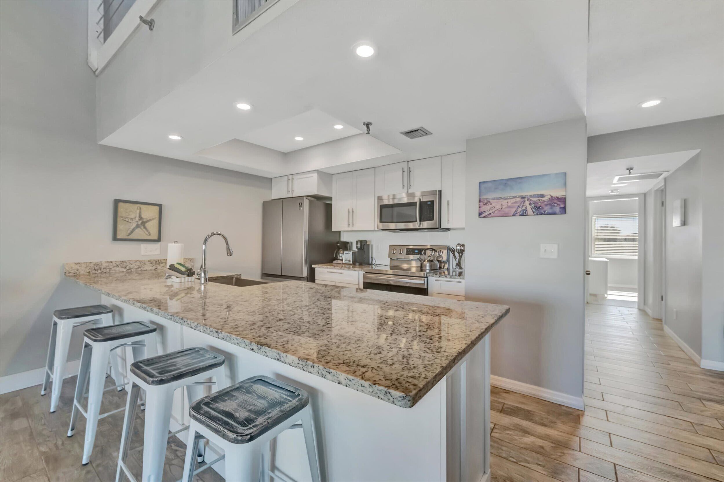 A modern kitchen with a granite island, white bar stools, stainless steel appliances, and a high ceiling with recessed lighting.