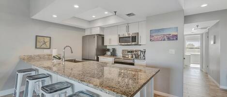 A modern kitchen with a granite island, white bar stools, stainless steel appliances, and a high ceiling with recessed lighting.