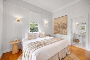 A bedroom with a large bed, two wall-mounted lights, a window, a painting, and a bathroom visible through an open door. - Paddy's Place - 6 Paddy's Crt, Bangalow (Byron)