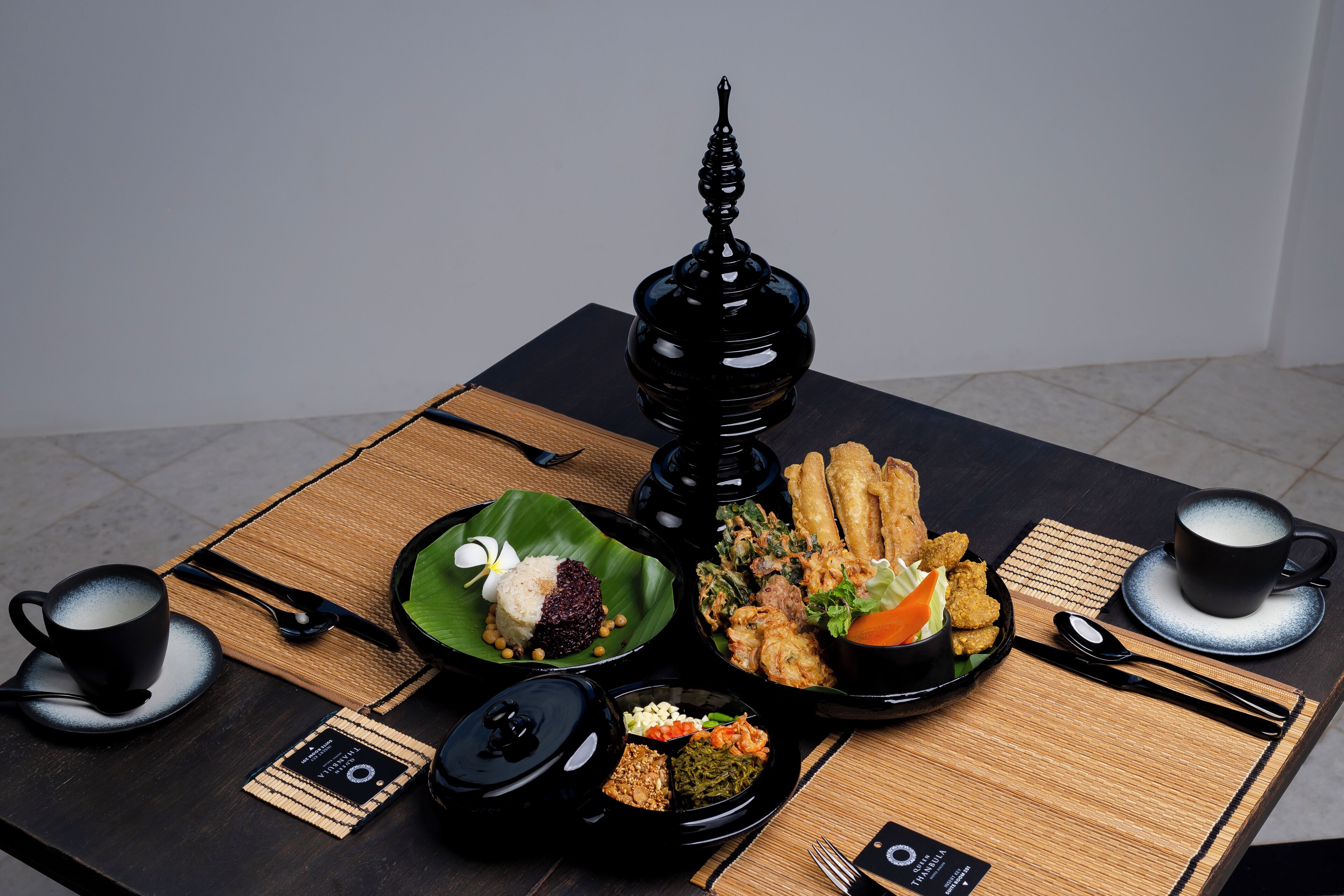 A table set with various dishes, including rice, vegetables, and fried items, along with black cups and chopsticks.