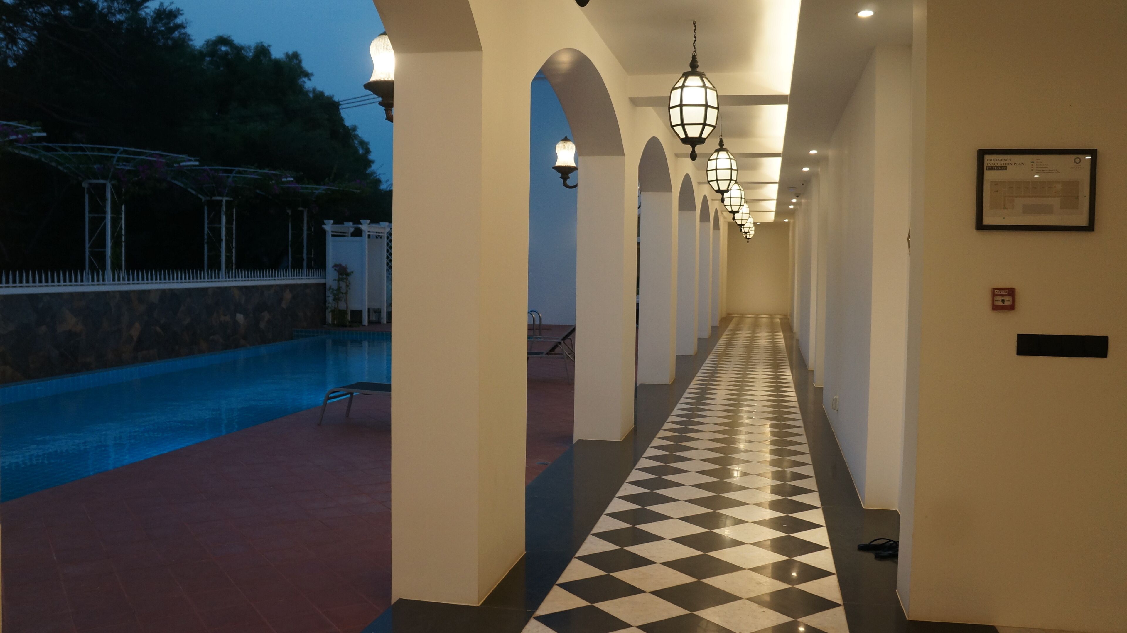 Un pasillo de hotel con piso a cuadros, columnas arqueadas y una zona de piscina visible a la izquierda.