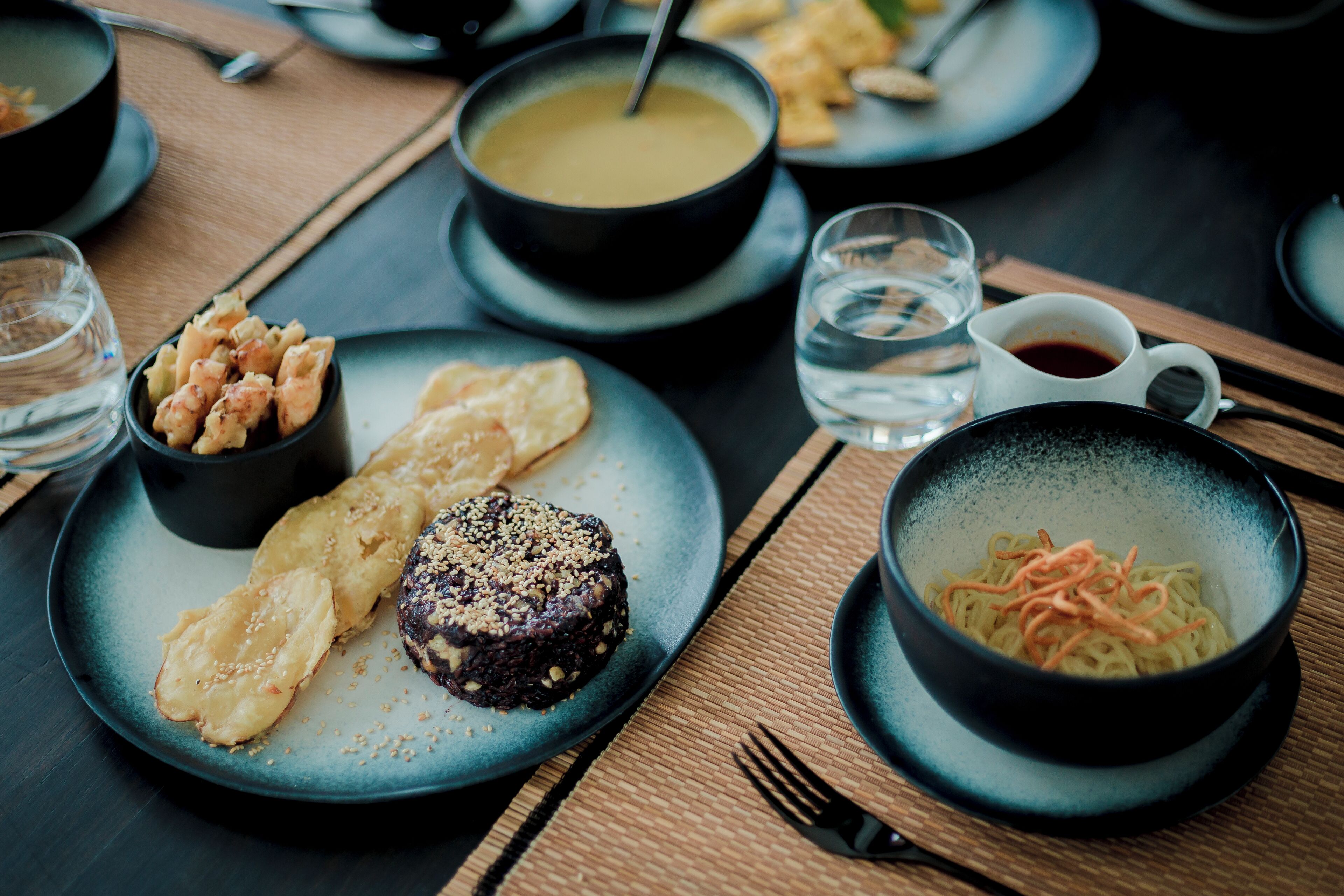 Una mesa de comedor con diversos platos, incluyendo un tazón de sopa, un plato con fideos y vegetales, y un postre.