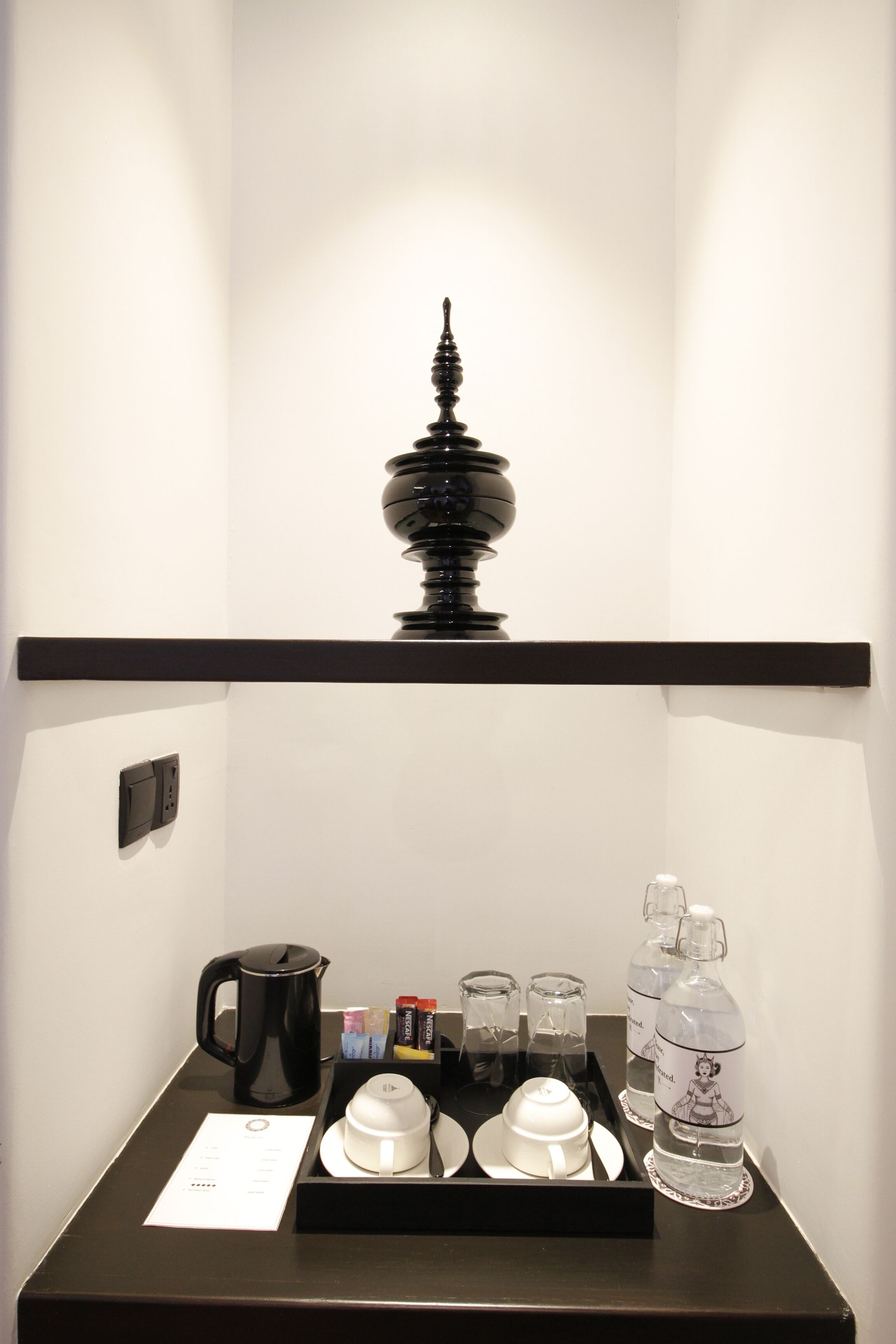 Ein Hotelzimmer mit Minibar, einem schwarzen dekorativen Gegenstand an der Wand sowie einem Tablett mit Tassen, einem Wasserkocher und Flaschen.