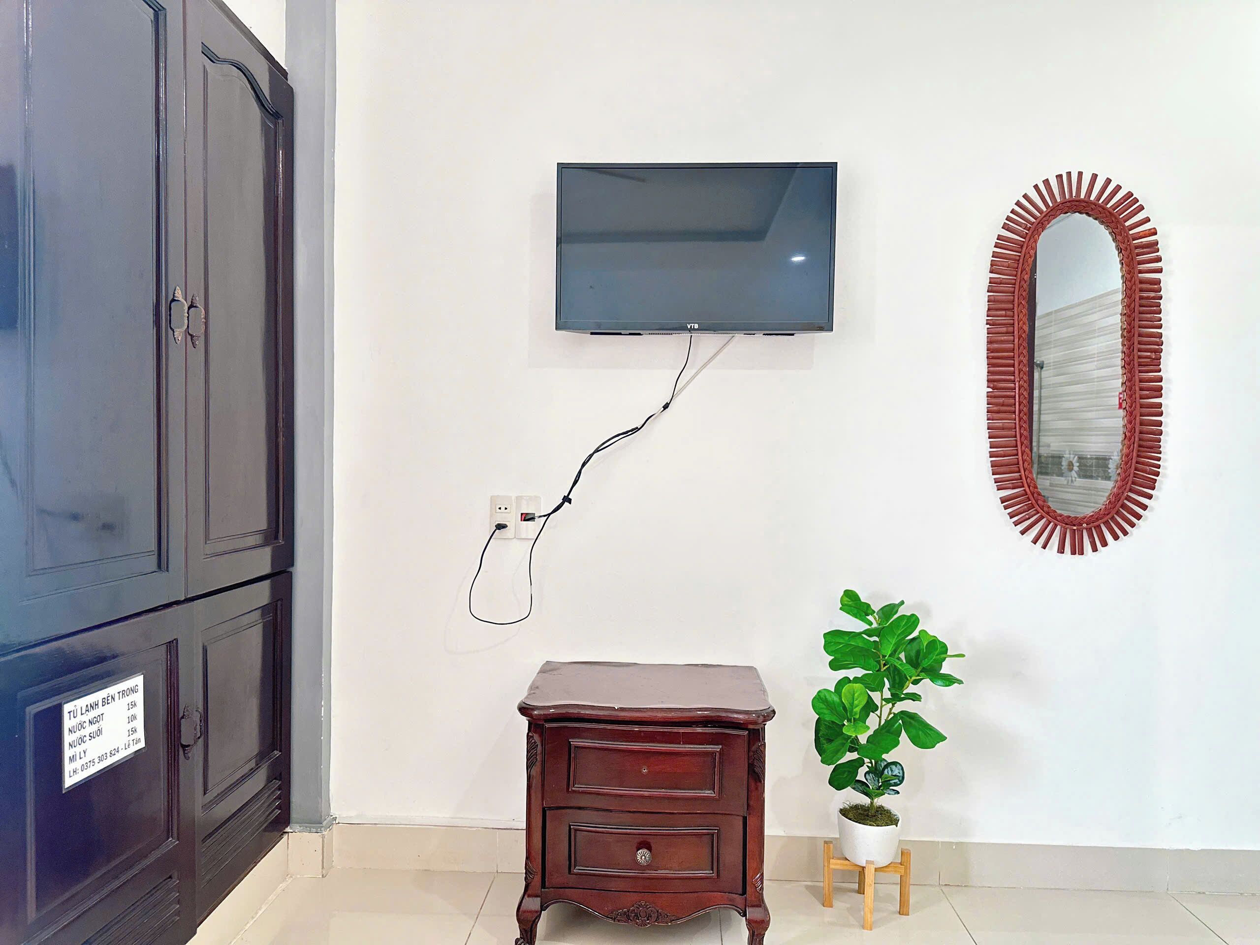 A room with a dark wooden wardrobe, a small wooden dresser, a television mounted on the wall, and a mirror with a red frame.