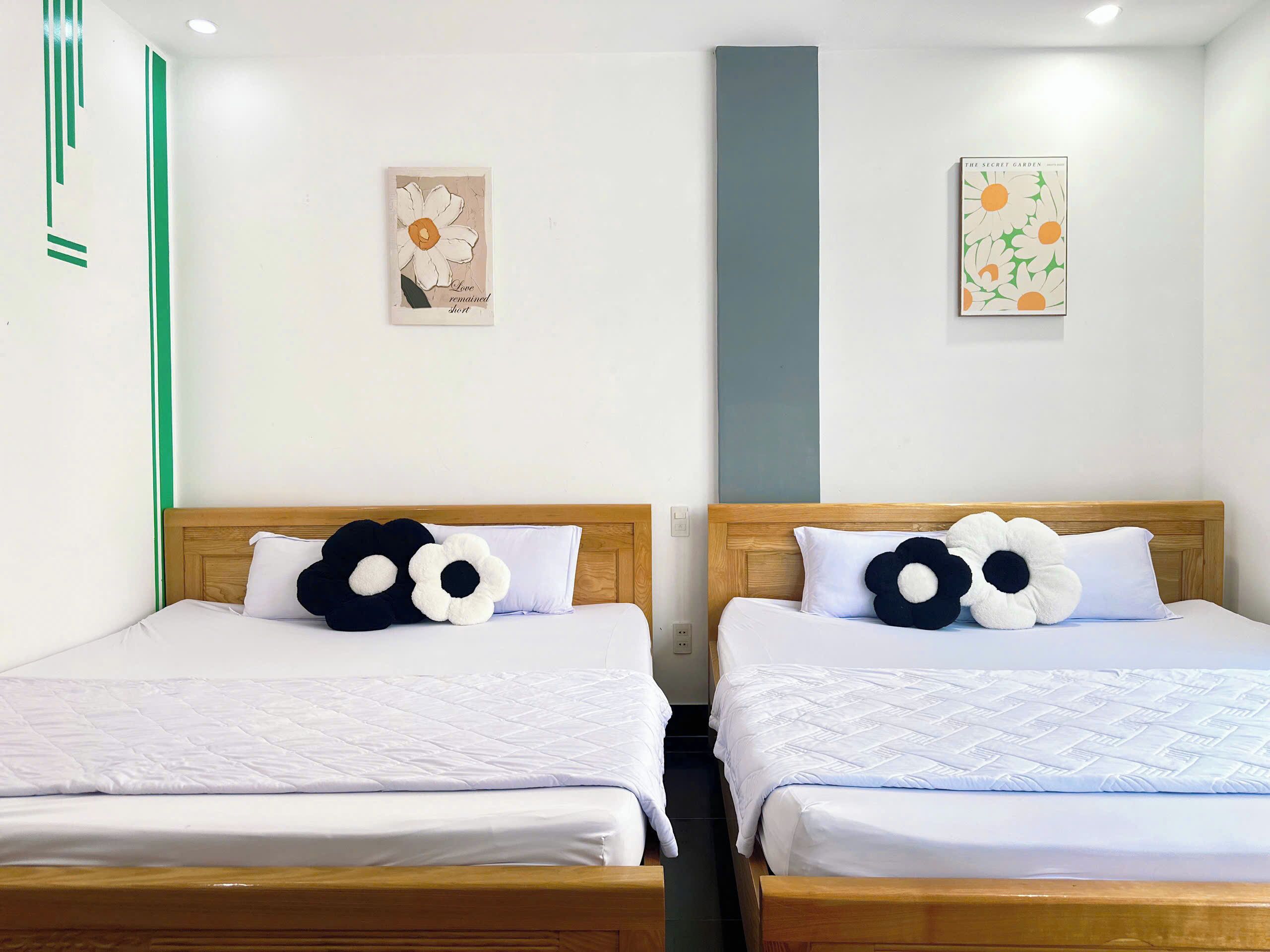 Two beds with white bedding and large flower-shaped pillows, a wooden headboard, and a wall with two framed pictures.