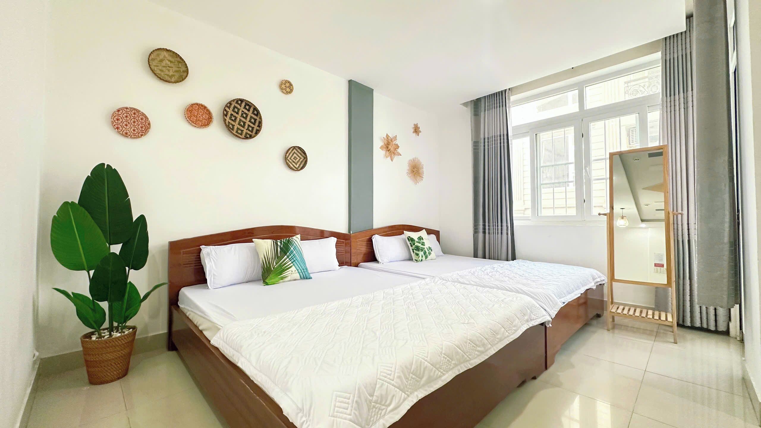 A hotel room with two beds, a large potted plant, decorative wall art, and a window with curtains.