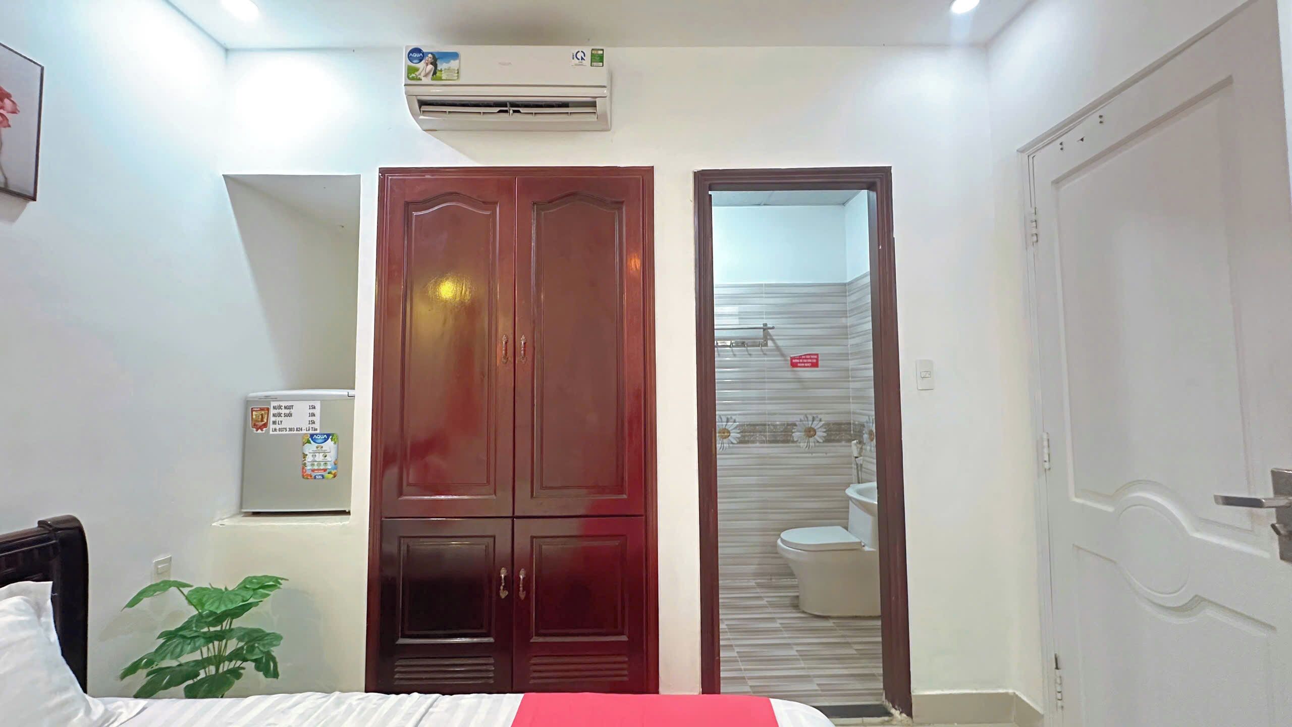 A bedroom with a bed, a wardrobe, an air conditioner, a refrigerator, and a bathroom visible through an open door.