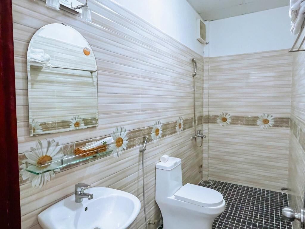 A bathroom with a white sink, toilet, and shower. There is a glass shelf with a flower decoration.