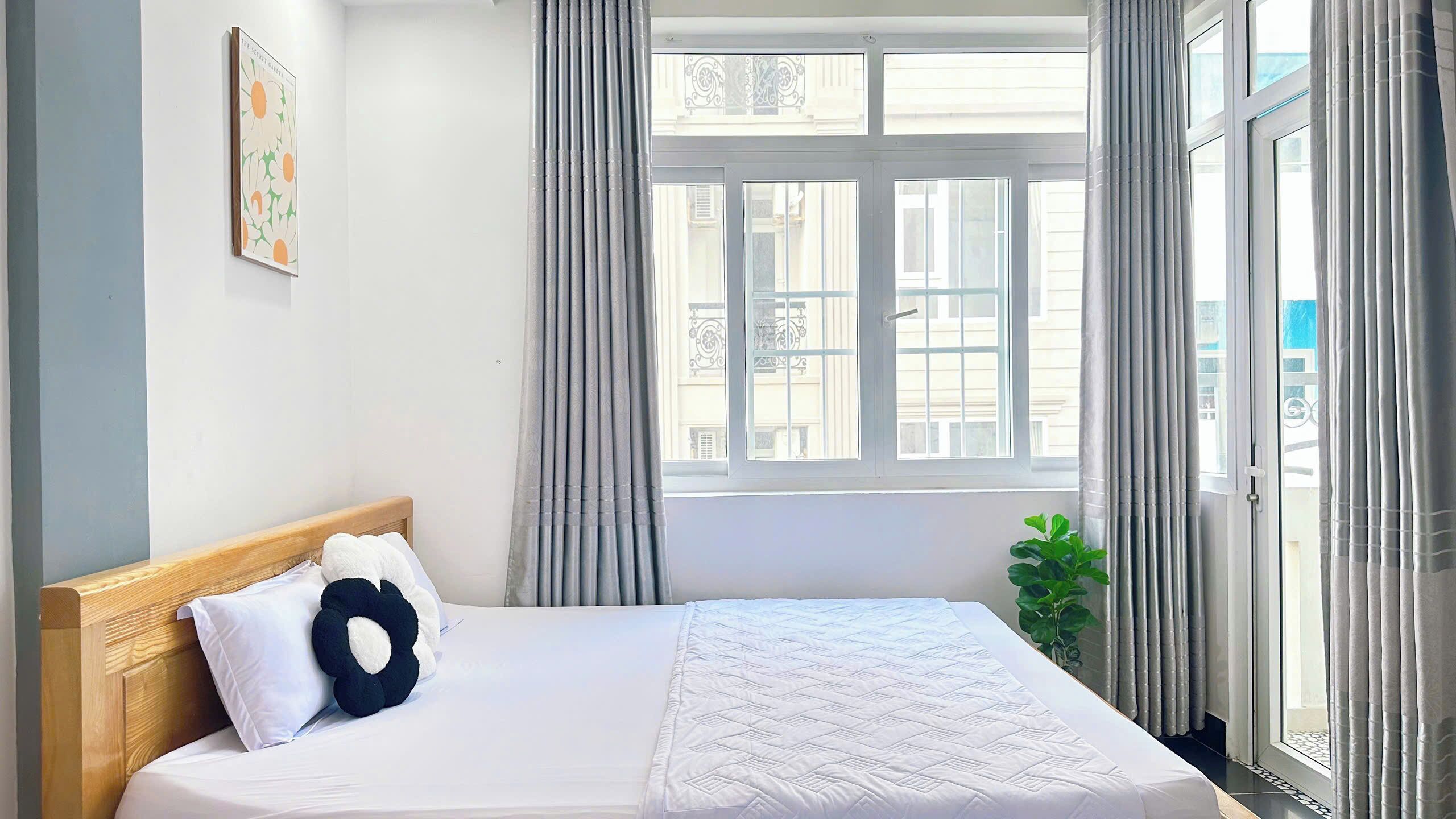 Hotellihuone, jossa on sänky, ikkuna, jossa on verhot, huonekasvi ja taulu seinällä.