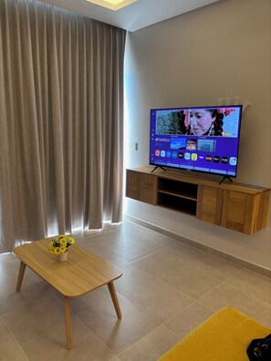 A living room with a flat-screen TV on a wooden stand, a small wooden table with a flower vase, and a yellow rug. - Elegant 2BR Marina-view condo (Crucecita)