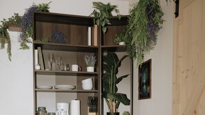 Un comedor con una mesa de madera, dos sillas, una estantería con plantas y una zona de cocina con tostadora y cafetera.