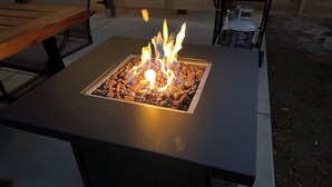 Un pozo de fuego cuadrado con leña encendida, rodeado por una mesa y un banco de hormigón.