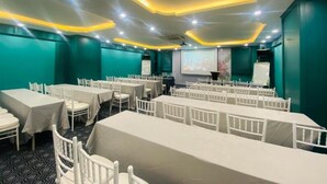 Un salón de conferencias con filas de mesas y sillas blancas, paredes color turquesa y una pantalla de proyector.