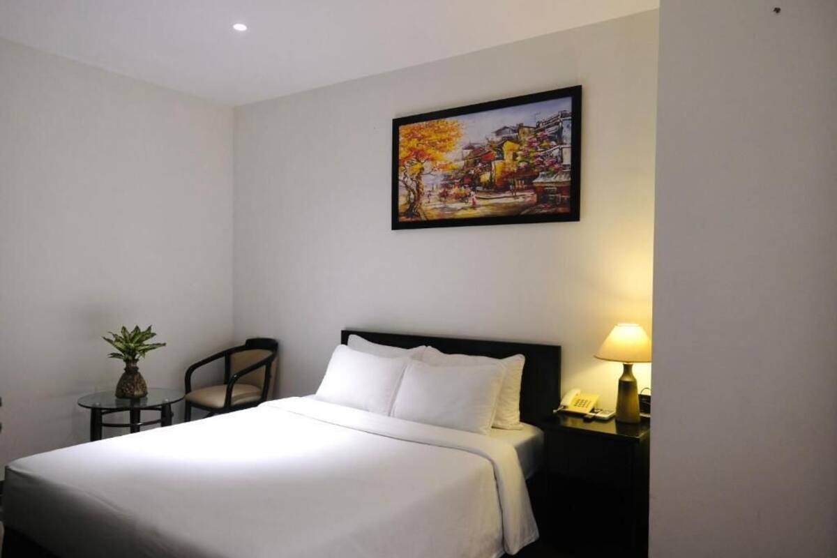 A hotel room with a bed, a small table with a plant, a chair, a nightstand with a lamp, and a painting on the wall.