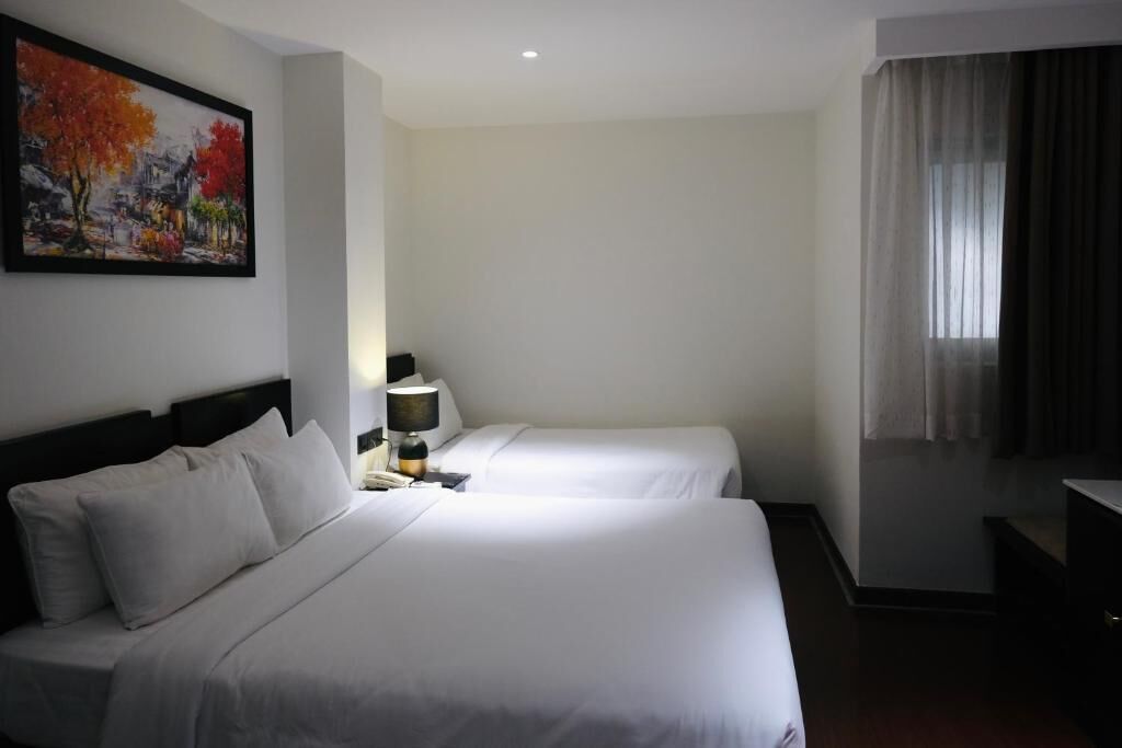 A hotel room with two single beds, a nightstand with a lamp, a painting on the wall, and a window with curtains.