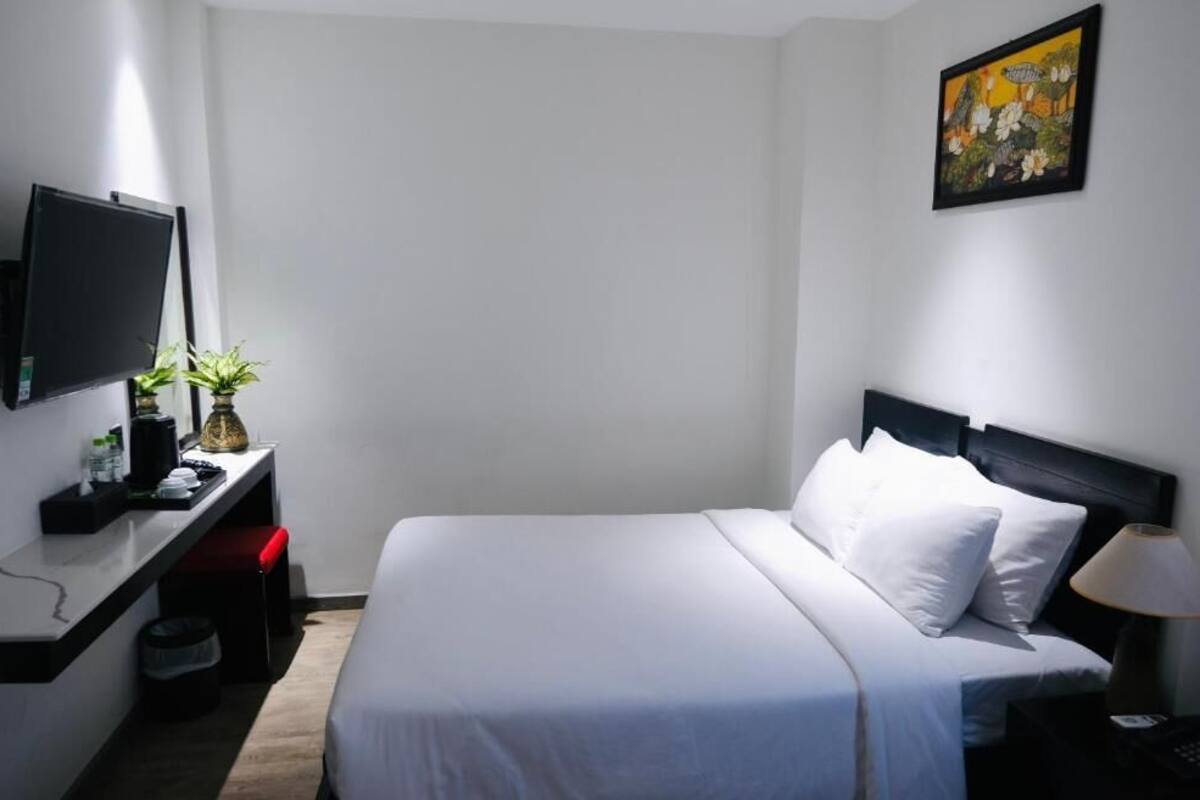 A hotel room with a bed, a desk with a TV, a plant, and a painting on the wall.