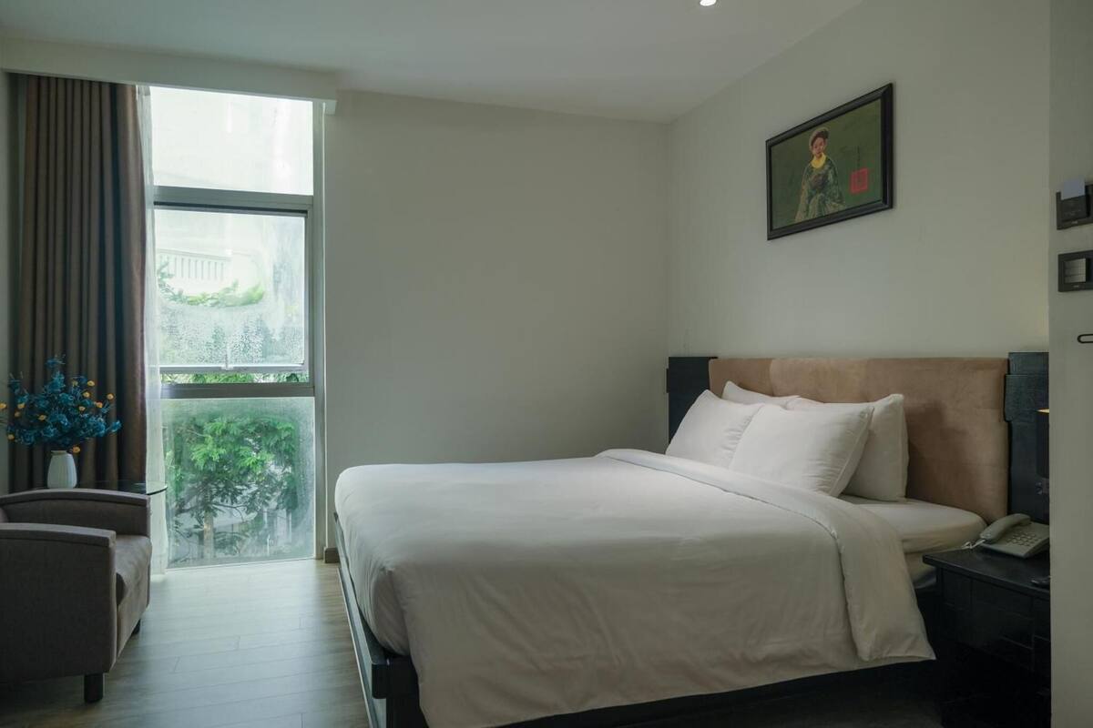 A hotel room with a large bed, a nightstand, a chair, and a window with a view of greenery.