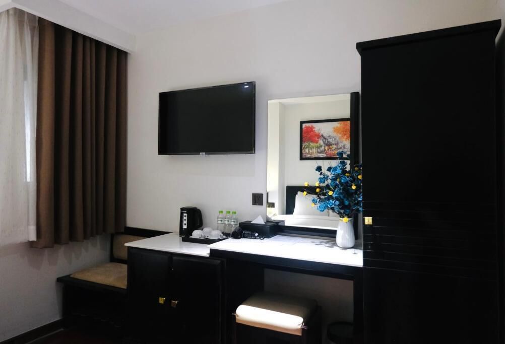 Habitación de hotel con televisión de pantalla plana en la pared, escritorio con cafetera, florero con flores y un mueble grande.