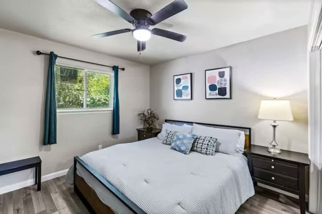 Een slaapkamer met een bed, een plafondventilator, een raam met gordijnen, een nachtkastje met een lamp, en twee ingekaderde foto’s aan de muur.