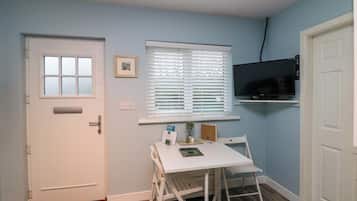 A small, well-lit room with a white door, a window with blinds, a mounted TV, a white table with a chair, and a small plant.
