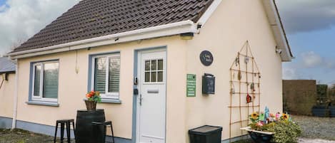 A small, single-story building with a tiled roof, a white door, and a small porch area with a stool and a barrel.