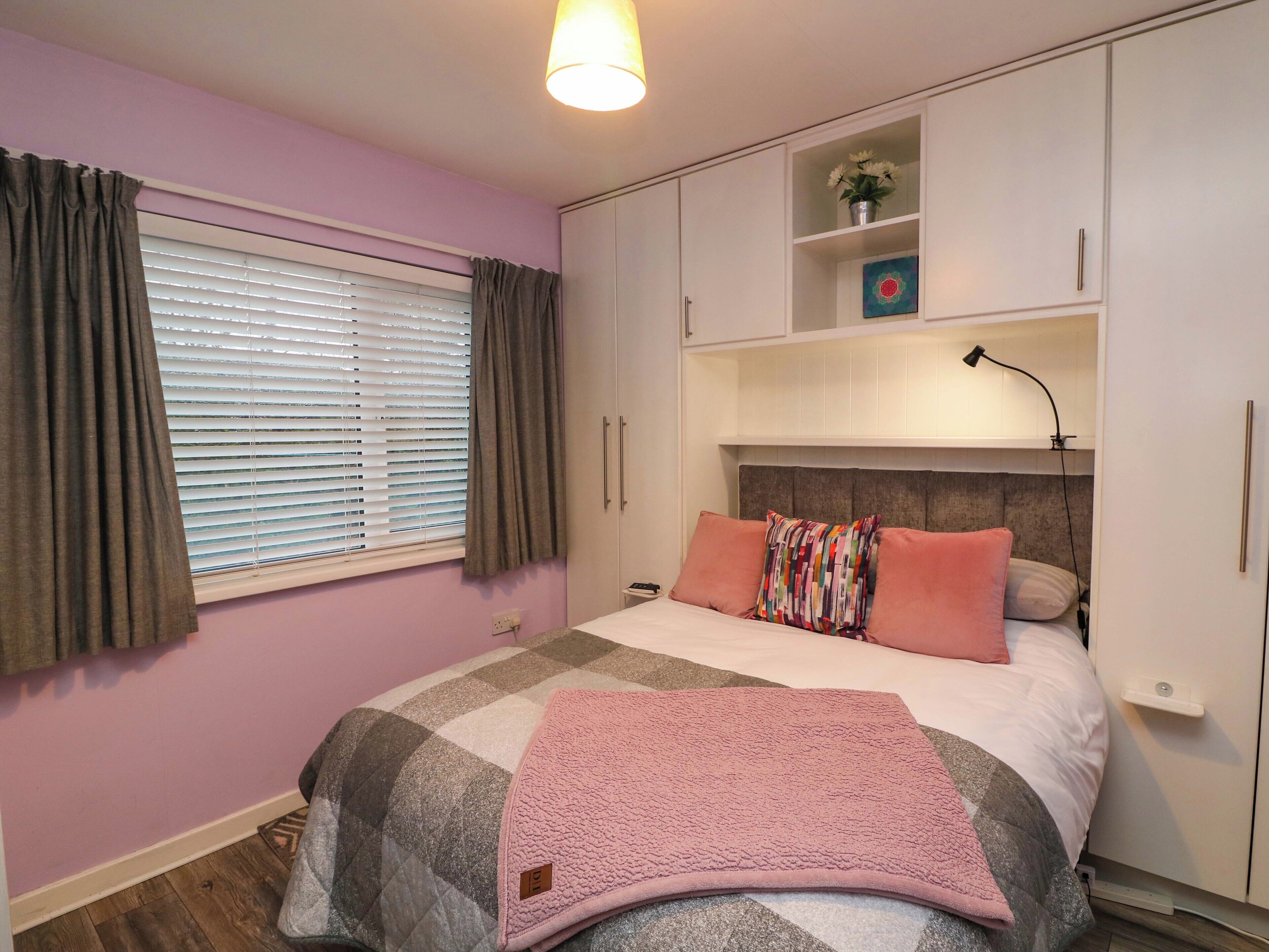 A bedroom with a bed, a bedside table, a lamp, a shelf with decorative items, and a window with curtains.