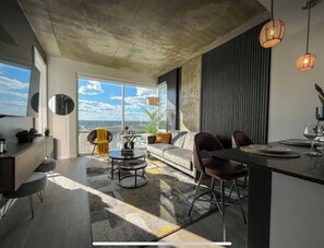 Ein modernes Wohnzimmer mit großem Fenster, einer Couch, einem Couchtisch und einem Essbereich mit Stühlen und einem Tisch.