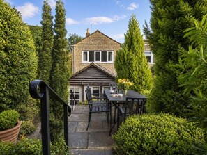 Ein Haus mit einer Steinfassade, einer Veranda mit Tisch und Stühlen und einem gepflegten Garten mit verschiedenen Sträuchern und Bäumen.