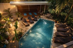 Una piscina rectangular con sillas de descanso y colchonetas de ejercicios, rodeada de plantas en macetas y un área de estar cubierta.