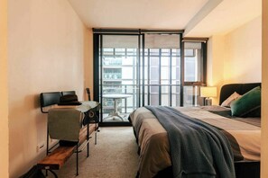Ein kleines Hotelzimmer mit Bett, Schreibtisch, Stuhl, einer Gitarre und einem Balkon mit Blick auf die Stadt.