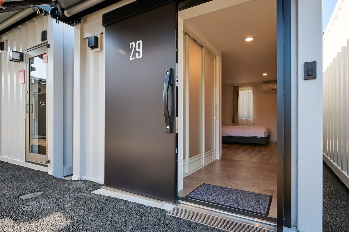 A modern hotel entrance with a dark door marked "29", a glass door to the left, and a bed visible inside.