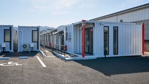 Uma fileira de edifícios modulares brancos com detalhes em vermelho, cada um com uma porta e janelas, e uma rampa acessível a cadeiras de rodas que leva à entrada.