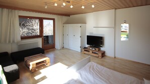 Quarto com uma cama, televisão, mesa de centro de madeira, sofá, janela com cortinas e porta.