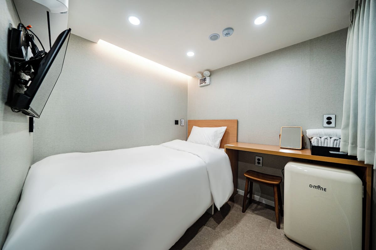 Ein Hotelzimmer mit einem Einzelbett, einem Schreibtisch mit Stuhl, einem kleinen Kühlschrank und einem Wandheizgerät.