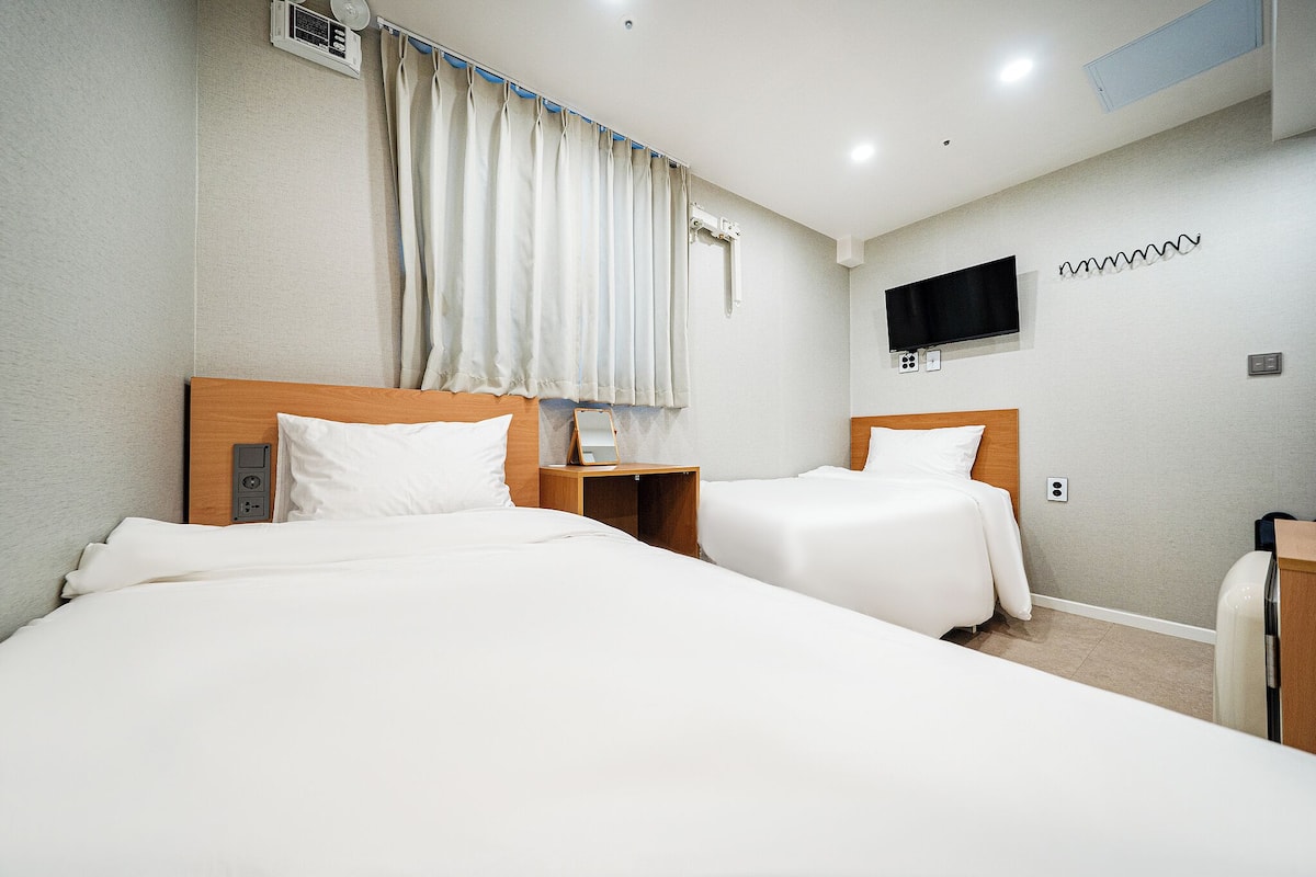 Ein Hotelzimmer mit zwei Betten, einem an der Wand befestigten Fernseher und einem Fenster mit Vorhängen.