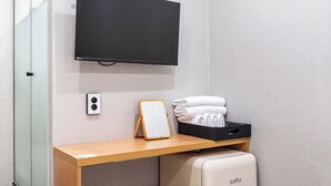 Una habitación de hotel con televisor de pantalla plana montado en la pared, un escritorio de madera con una pizarra, un caja de almacenamiento negra con toallas y una maleta blanca.