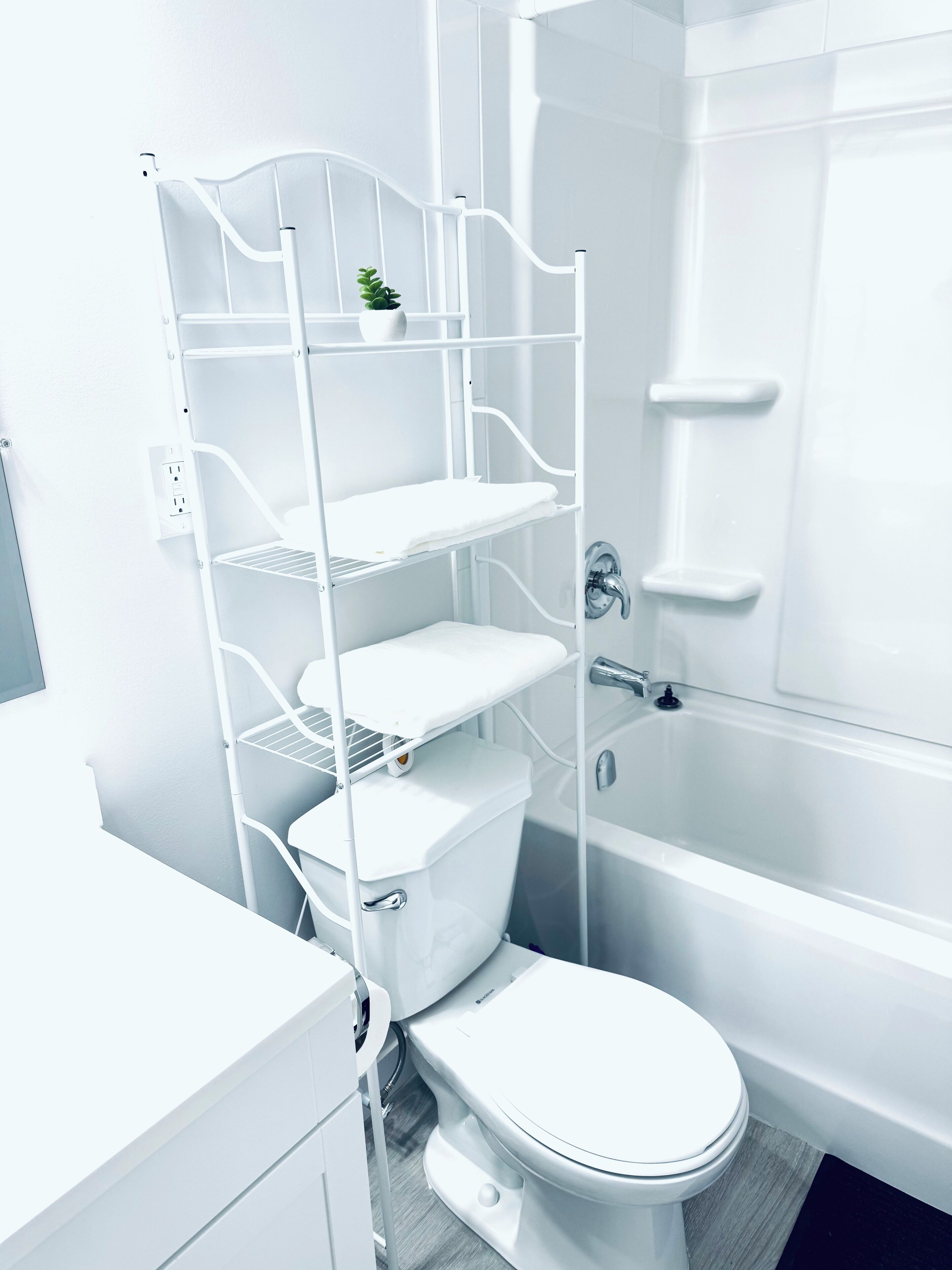 A modern bathroom with a white toilet, a glass-enclosed shower, and a white metal shelf unit with a small plant.