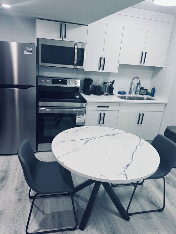 Een moderne keuken met witte kastjes, een roestvrijstalen koelkast, een inbouwoven en een magnetron. Er staat een rond marmeren tafel met twee zwarte stoelen.