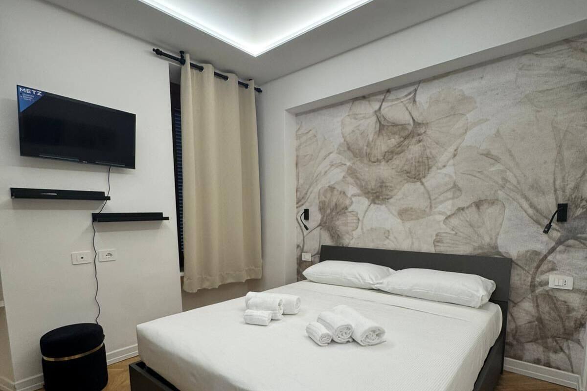 A modern bedroom with a bed, a mounted TV, a wall-mounted power outlet, and a small black ottoman.