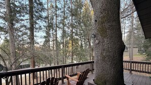 A wooden deck with two Adirondack chairs and a black bowl, surrounded by tall trees.