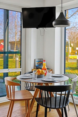 A dining area with a round table, chairs, a television mounted on the wall, and a window with a view of trees and a building. - Apartment am See by Interhome (Lembruch)