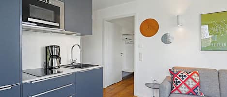 Eine moderne Küche mit Kaffeemaschine, Mikrowelle und Spüle. Im Wohnbereich befindet sich ein graues Sofa mit einem gemusterten Kissen und ein kleiner runder Tisch.