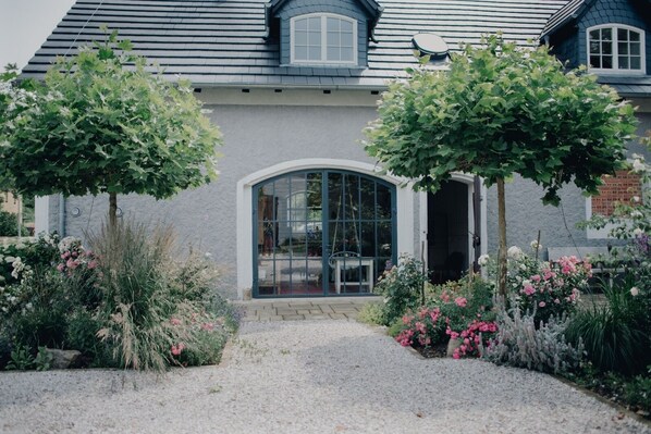 Une maison avec une allée gravillonnée, une grande entrée en arc de cercle et un jardin agrémenté de diverses plantes et fleurs.