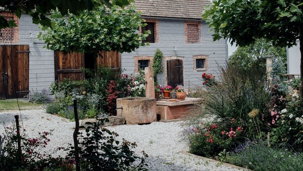 A rustic building with a gravel courtyard, surrounded by lush greenery and flowering plants. - Quiet apartment above the Bobertal valley in the foothills of the Giant Mountains (Tarczyn)