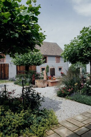 Un bâtiment rustique doté d’une cour en gravier, entouré d’une végétation luxuriante et de plantes en fleurs.
