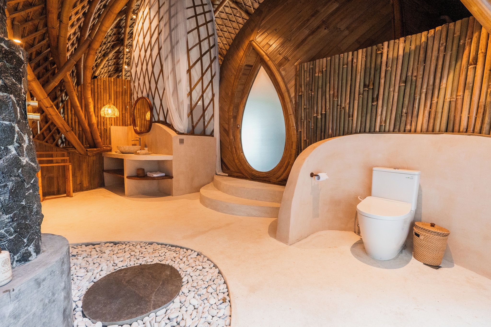 An interior with a stone wall, a wooden ceiling, a round mirror, a white toilet, and a woven basket.