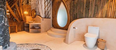 An interior with a stone wall, a wooden ceiling, a round mirror, a white toilet, and a woven basket.