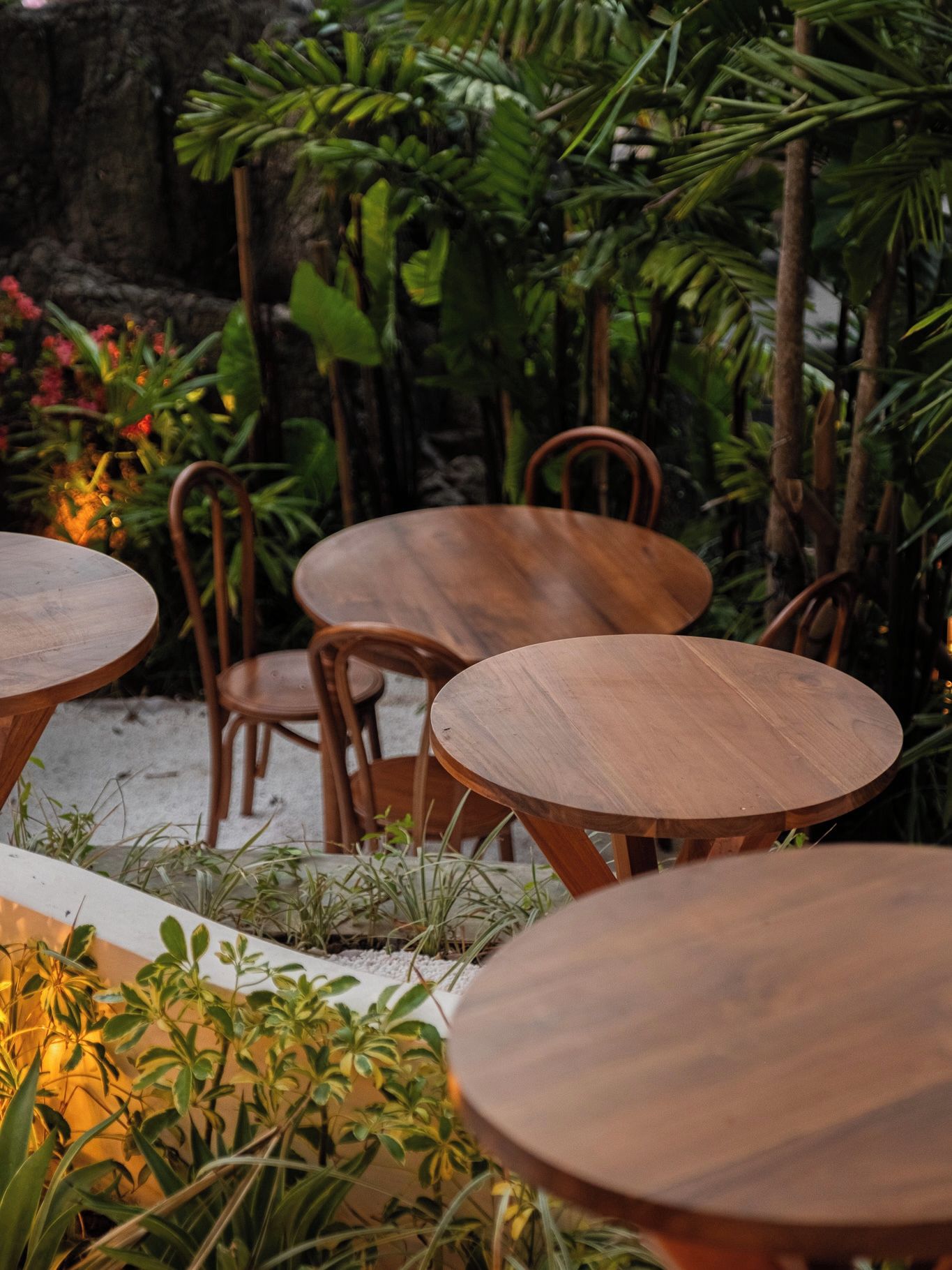 A wooden table and chairs set in an outdoor area with lush greenery and a stone wall in the background.